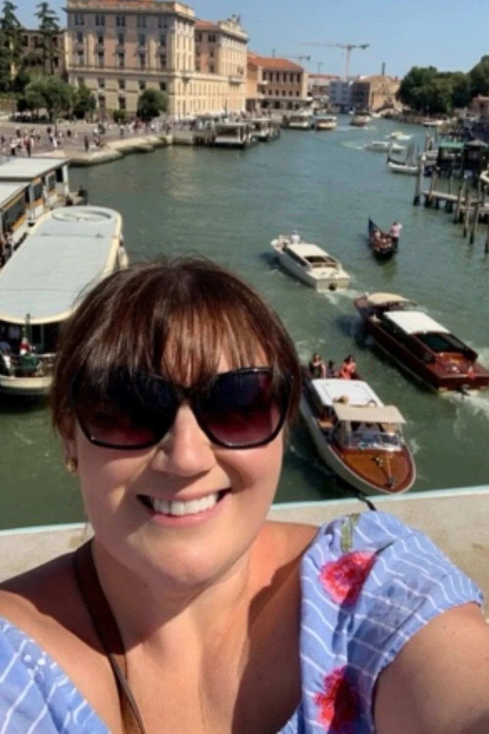 Gemma Lawrence taking a selfie on a bridge overlooking a canal in Venice, Italy. Below, several boats, including water taxis and gondolas, navigate the canal, with historic buildings lining the waterway in the background.