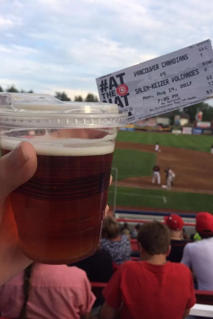 Foreground shows a clear plastic cup of beer with a baseball game ticket resting on top, highlighting a game between the Vancouver Canadians 