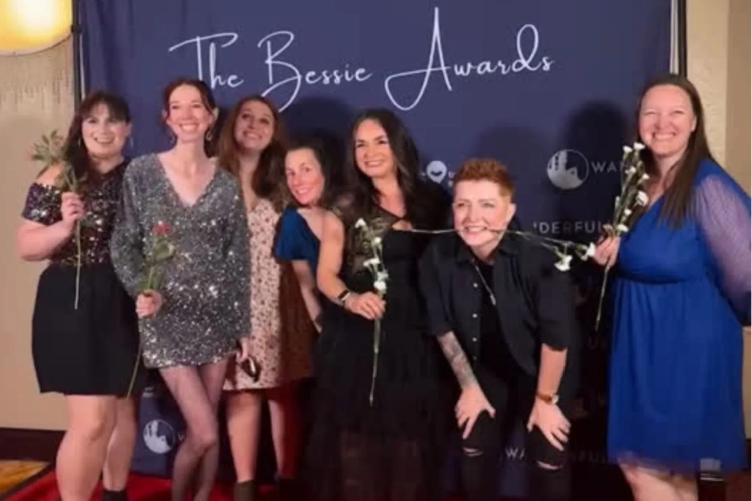 Gemma Lawrence, Hannah Dixon and five other women posing on the red carpet at the Bessie Awards, some holding white roses, in front of a backdrop with 'The Bessie Awards' text.