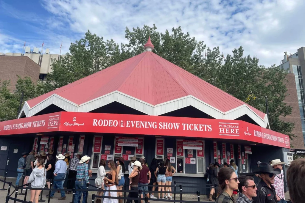 Crowd gathers outside the Calgary Stampede ticket booth under a red and white tent-style roof, with signs reading “Rodeo & Evening Show Tickets” and prices for events prominently displayed.