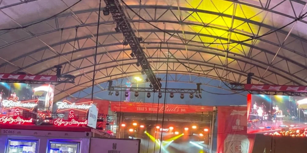 Inside a crowded Nashville North with people seated at tables and standing near the bar, watching a performance on stage. The tent is filled with neon signs and large video screens showing the performer on stage. The atmosphere is energetic, with people enjoying the live music and vibrant surroundings.