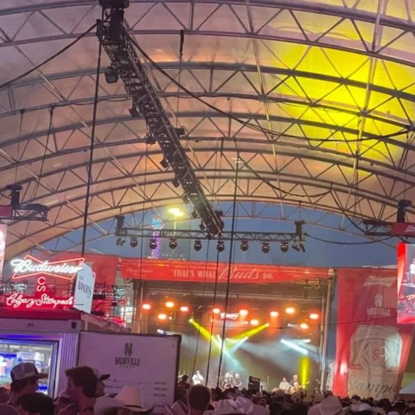 Inside a crowded Nashville North with people seated at tables and standing near the bar, watching a performance on stage. The tent is filled with neon signs and large video screens showing the performer on stage. The atmosphere is energetic, with people enjoying the live music and vibrant surroundings.
