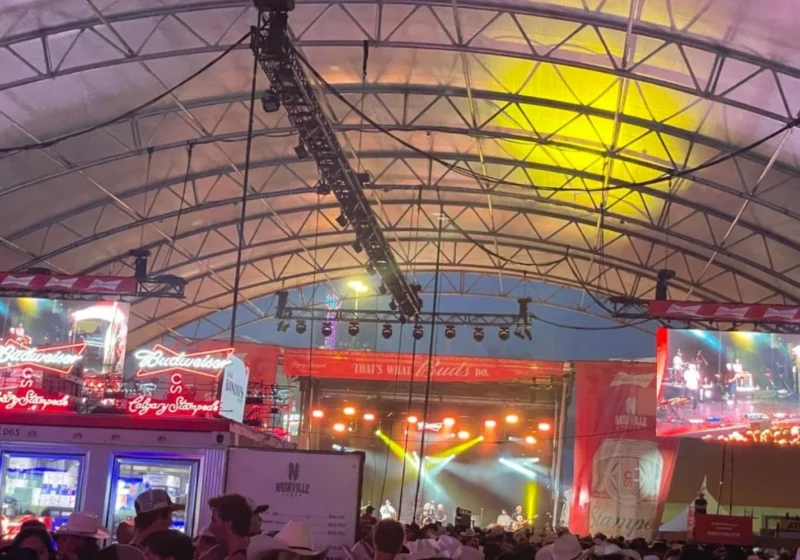Inside a crowded Nashville North with people seated at tables and standing near the bar, watching a performance on stage. The tent is filled with neon signs and large video screens showing the performer on stage. The atmosphere is energetic, with people enjoying the live music and vibrant surroundings.