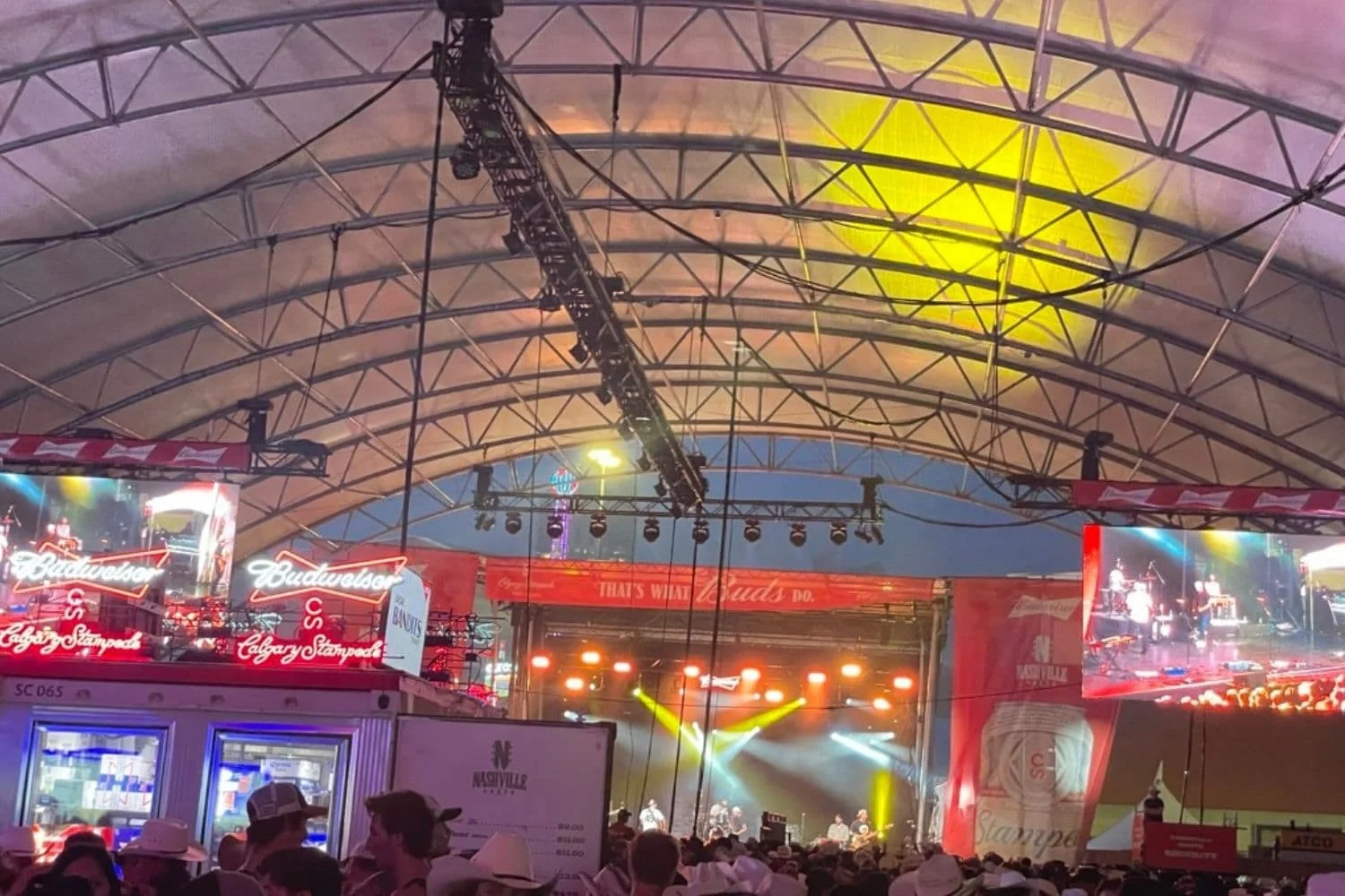 Inside a crowded Nashville North with people seated at tables and standing near the bar, watching a performance on stage. The tent is filled with neon signs and large video screens showing the performer on stage. The atmosphere is energetic, with people enjoying the live music and vibrant surroundings.