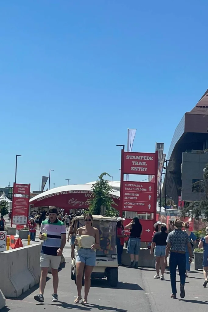 Crowds enter the Calgary Stampede grounds through the Stampede Trail Entry, with a red sign showing directions for tickets and services, and a white canopy in the background reading “Welcome to the Greatest Outdoor Show.”
