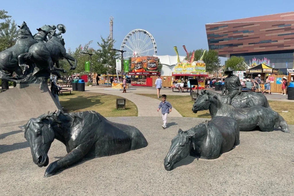 A daytime scene of Stampede Park with a bronze statue of a cowboy riding a bucking horse, surrounded by other horse sculptures. A child is walking near the statues, and fair booths with food and games are visible in the background, along with a large Ferris wheel