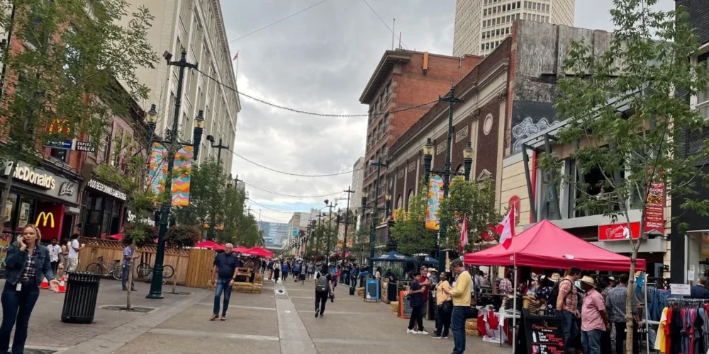 A lively street scene on Stephens Avenue in Calgary during Calgary Stampede, featuring people walking between shops and outdoor stalls under a cloudy sky. A McDonald’s and various local businesses are visible along the street.