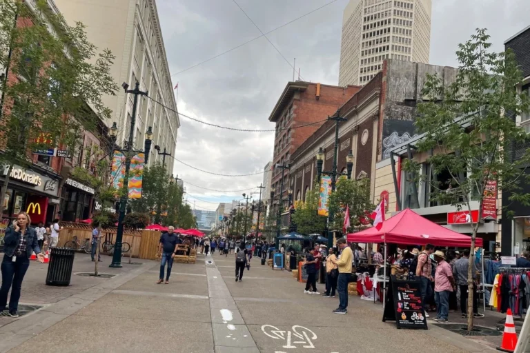 A lively street scene on Stephens Avenue in Calgary during Calgary Stampede, featuring people walking between shops and outdoor stalls under a cloudy sky. A McDonald&rsquo;s and various local businesses are visible along the street.