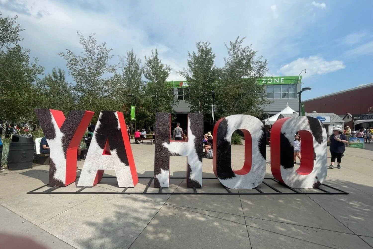 Large, freestanding letters spelling out "YAHOO" with a cow-print design, set against an outdoor backdrop with trees and a few people sitting at picnic tables on Stampede Park.