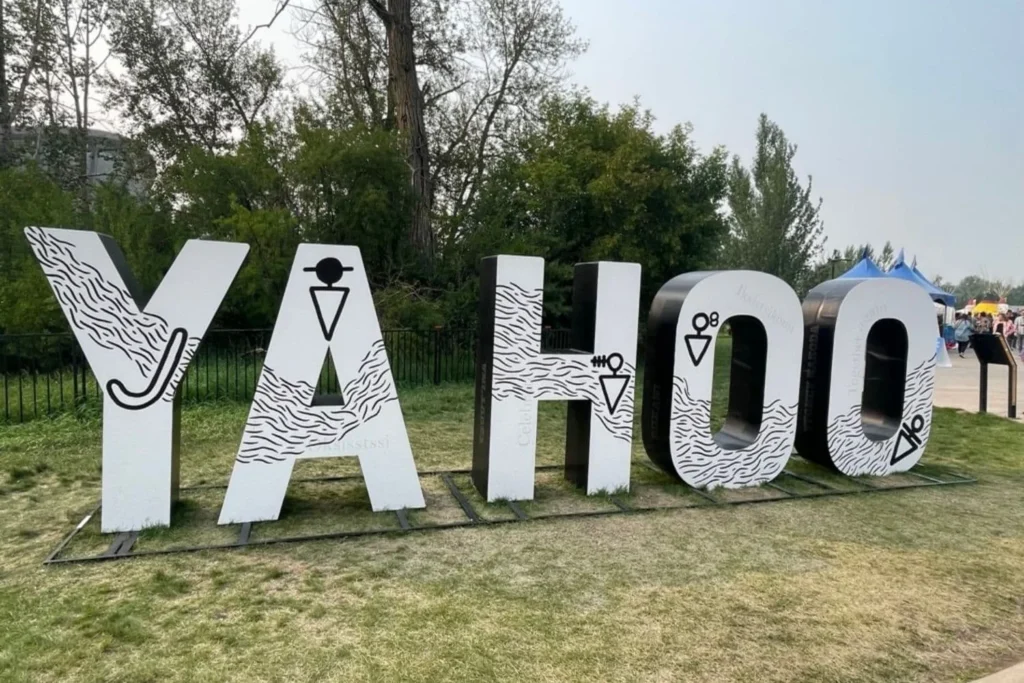 A large outdoor sign spelling "YAHOO" in white letters with black outlines and Western-themed designs, set up on a grassy area with trees in the background at the Elbow River Camp in Calgary.