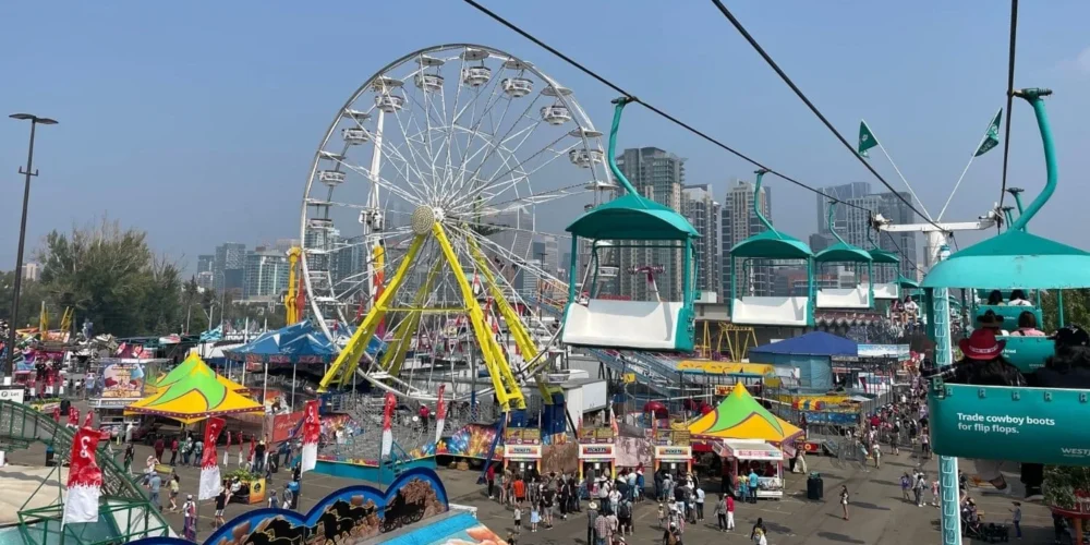 A colorful view of the Calgary Stampede midway, showcasing a large Ferris wheel, various food stands, and the westjet skyride with people riding in teal seats.