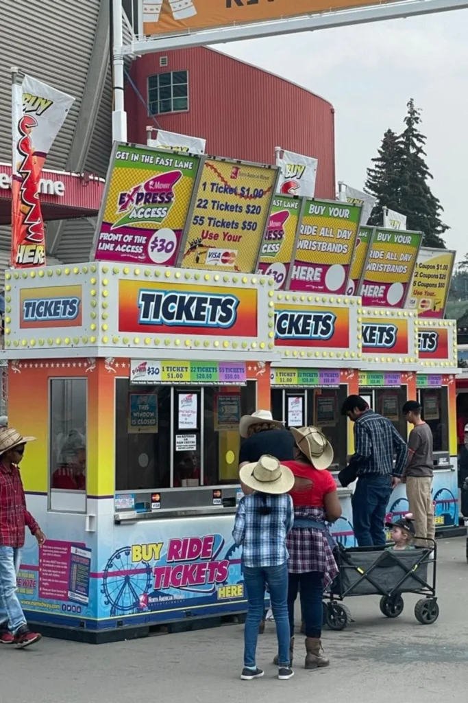 A group of people, many wearing cowboy hats, line up at colorful ticket booths at a midway. The booths are brightly decorated with signs advertising ride tickets, wristbands, and express access, with a large sign overhead indicating the location.