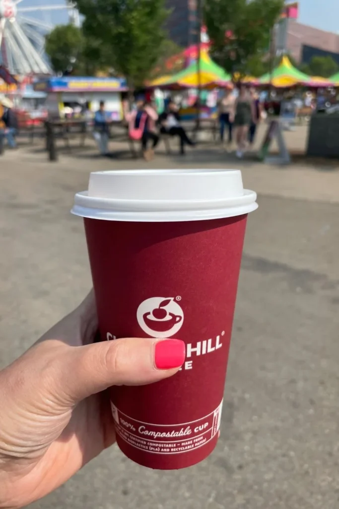 A hand holding a red paper coffee cup from Good Earth Coffeehouse at the Calgary Stampede, with a carnival area visible in the background.