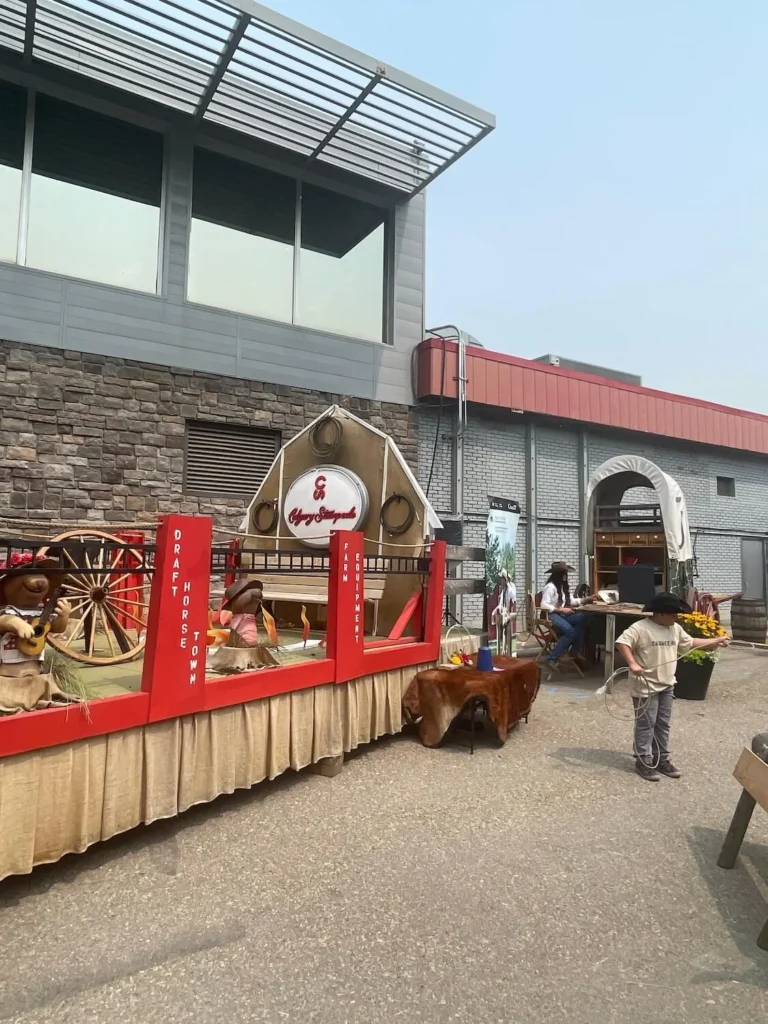 The entrance area of Draft Horse Town at the Calgary Stampede, featuring a display with small cowboy-themed stuffed toys in a red and white exhibit labeled "Draft Horse Town Farm Equipment." A young child in a cowboy hat is practicing roping in the foreground.