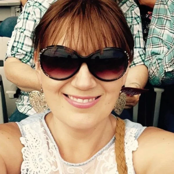 Gemma Lawrence taking a selfie while seated in a stadium, possibly at the Calgary Stampede. She is wearing sunglasses and a white and denim lace top, with others in cowboy attire seated behind her.