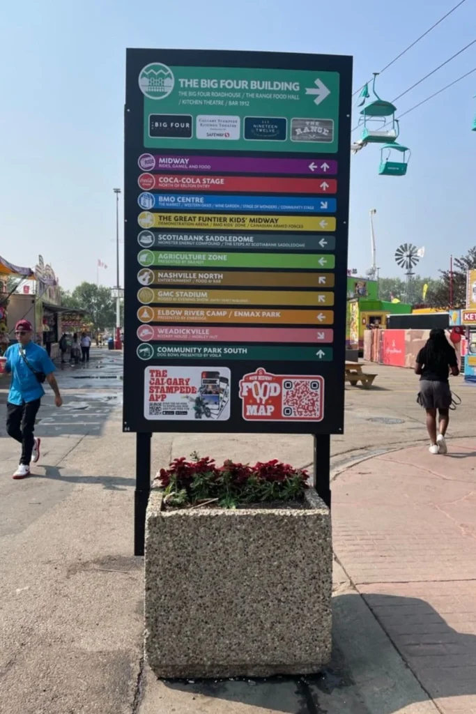 A close-up of a directional sign at a fairground, listing various locations like the Midway, Coca-Cola Stage, and Nashville North. A few people walk by in the background, and the sign promotes the Calgary Stampede app with a QR code.