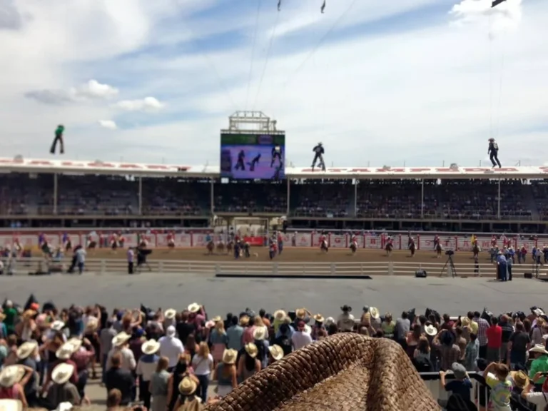 Calgary Stampede rodeo event with cowboys on horseback arriving in the arena, with a crowd in the stands watching. A large screen displays the event overhead.