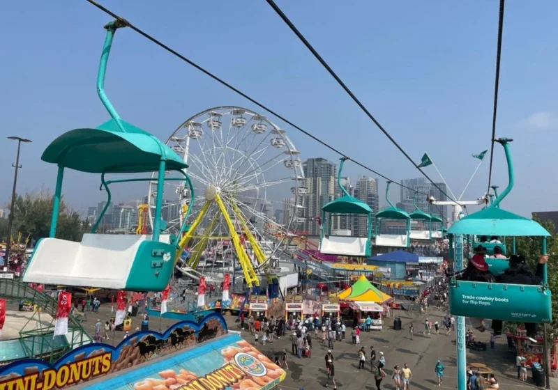 A view from a WestJet Skyride chair lift at Stampede Midway, looking down at the crowds and colorful rides below, including a Ferris wheel.