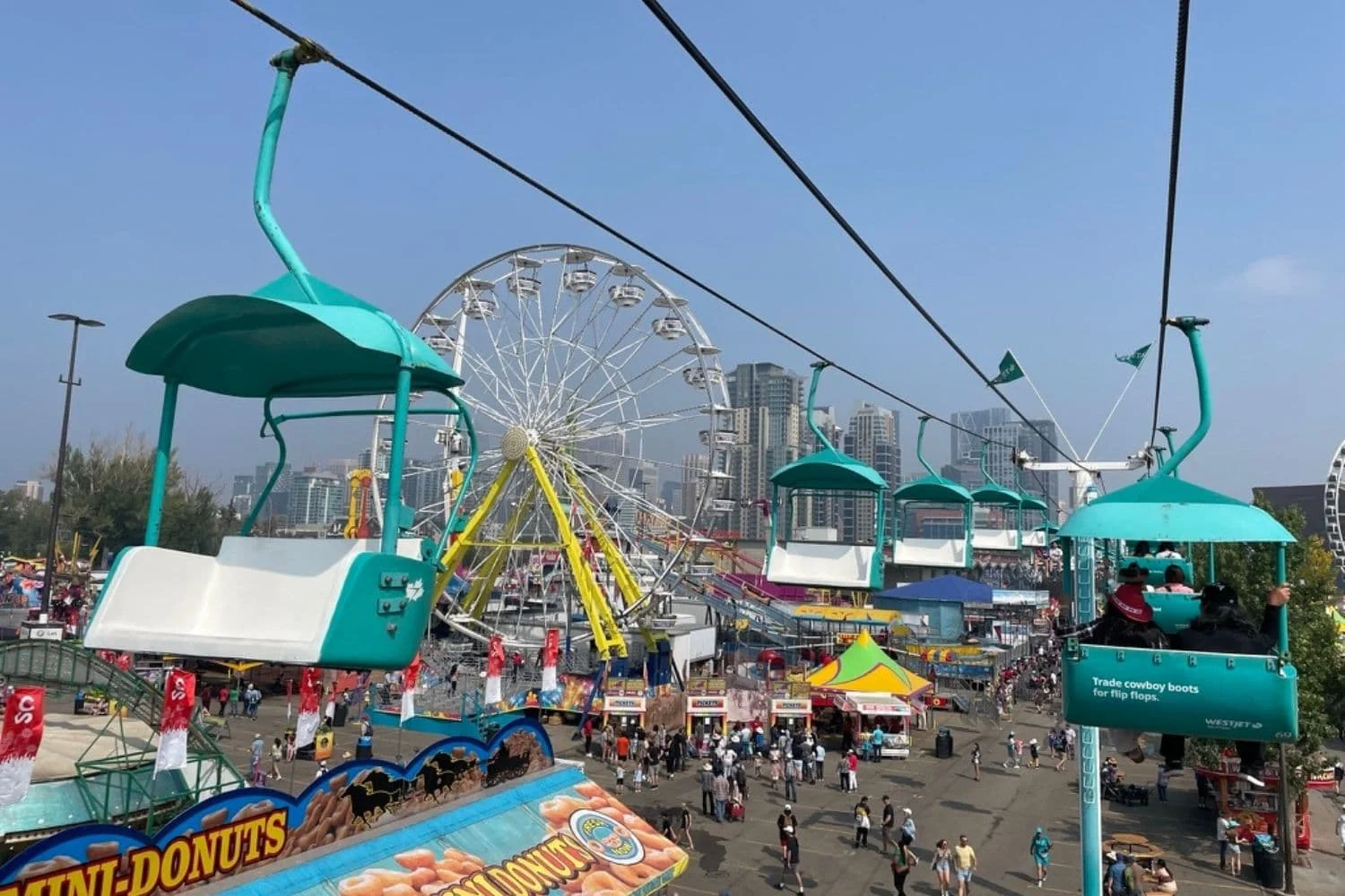 A view from a WestJet Skyride chair lift at Stampede Midway, looking down at the crowds and colorful rides below, including a Ferris wheel.