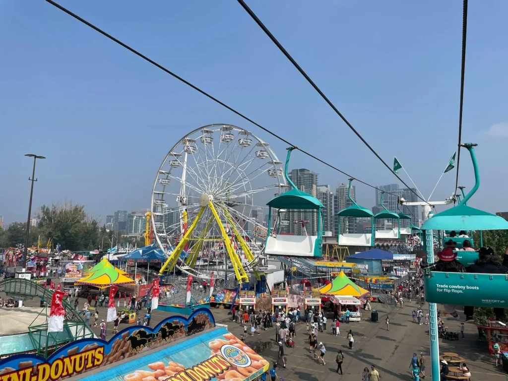 Another angle of the WestJet Skyride, showing a panoramic view of the Stampede Park Midway fairgrounds below, including the Ferris wheel and food stalls. The chairlifts carry people over the bustling scene, with tall buildings in the distance.