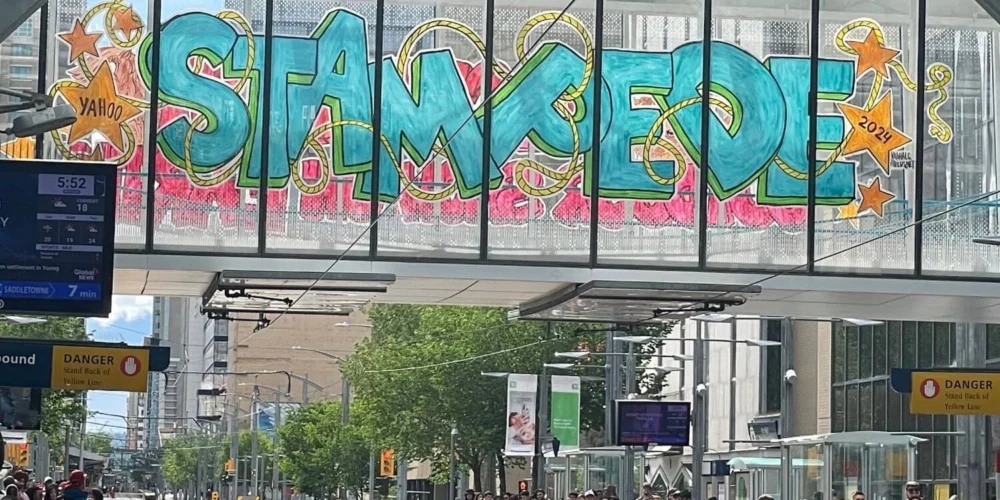 A large graffiti-style mural on a glass-covered walkway reads "STAMPEDE" in bright turquoise and pink colors, with stars and the year "2024" incorporated into the design. Below the walkway, people wait at a light rail station in the city of Calgary.
