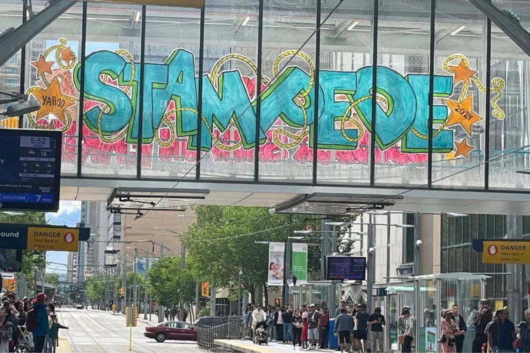 A large graffiti-style mural on a glass-covered walkway reads "STAMPEDE" in bright turquoise and pink colors, with stars and the year "2024" incorporated into the design. Below the walkway, people wait at a light rail station in the city of Calgary.