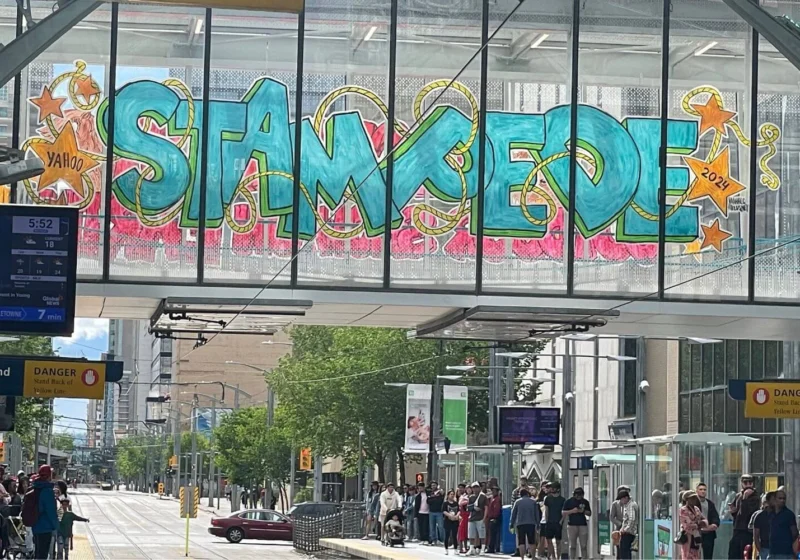 A large graffiti-style mural on a glass-covered walkway reads "STAMPEDE" in bright turquoise and pink colors, with stars and the year "2024" incorporated into the design. Below the walkway, people wait at a light rail station in the city of Calgary.