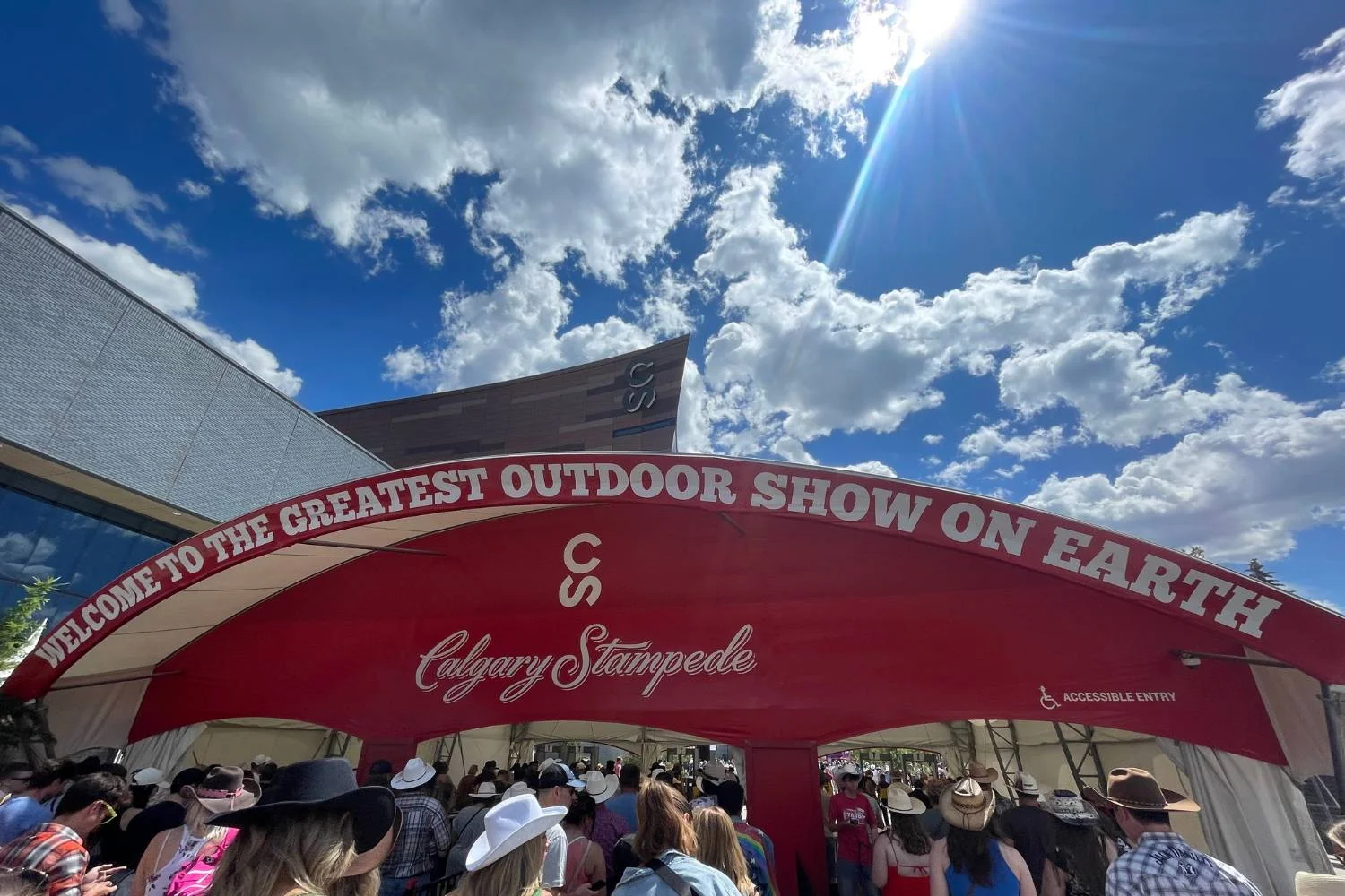 A large red entrance tent with white text reading "WELCOME TO THE GREATEST OUTDOOR SHOW ON EARTH" marks the entrance to the Calgary Stampede. A crowd of people wearing cowboy hats is walking through, with a bright blue sky and scattered clouds overhead.