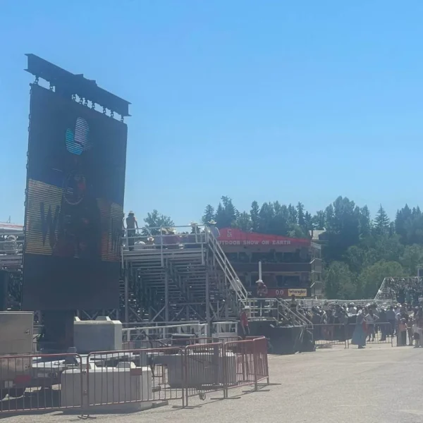 A large outdoor event setup for the afternoon rodeo at the Calgary Stampede, featuring a giant screen displaying a cowboy-themed image. Bleachers filled with spectators, a red "Outdoor Show of Earth" banner, and various event structures are visible under a bright blue sky.