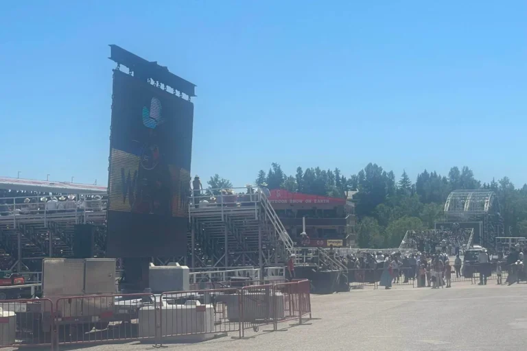 A large outdoor event setup for the afternoon rodeo at the Calgary Stampede, featuring a giant screen displaying a cowboy-themed image. Bleachers filled with spectators, a red "Outdoor Show of Earth" banner, and various event structures are visible under a bright blue sky.