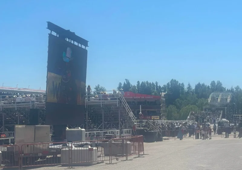 A large outdoor event setup for the afternoon rodeo at the Calgary Stampede, featuring a giant screen displaying a cowboy-themed image. Bleachers filled with spectators, a red "Outdoor Show of Earth" banner, and various event structures are visible under a bright blue sky.
