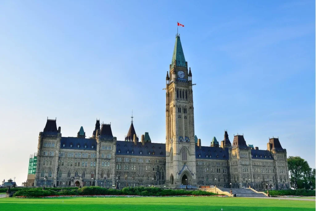 The iconic Parliament Hill building in Ottawa, Canada, with its Gothic Revival architecture, a Canadian flag atop the Peace Tower, and a lush green lawn in the foreground.