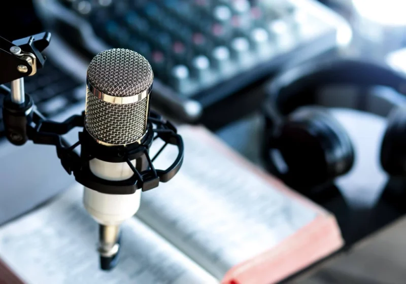 A close-up of a professional podcasting setup with a microphone, a pair of headphones, an open book, and a sound mixer in the background.