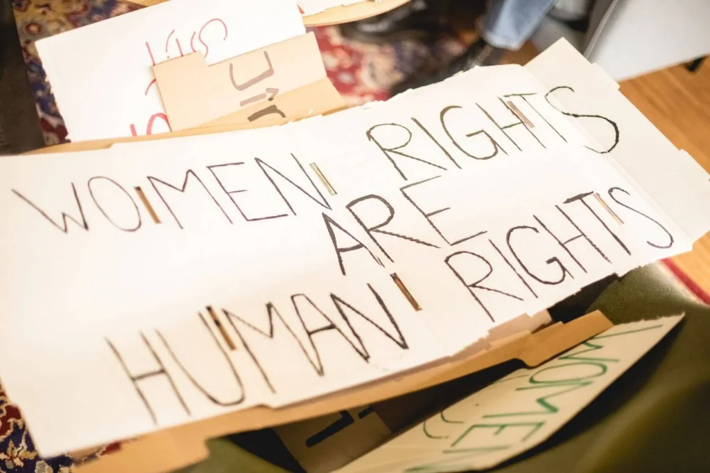 A handwritten protest sign on white cardboard reading "Women’s Rights Are Human Rights," placed among other protest materials. The sign is part of a collection on a table with a patterned carpet visible in the background.