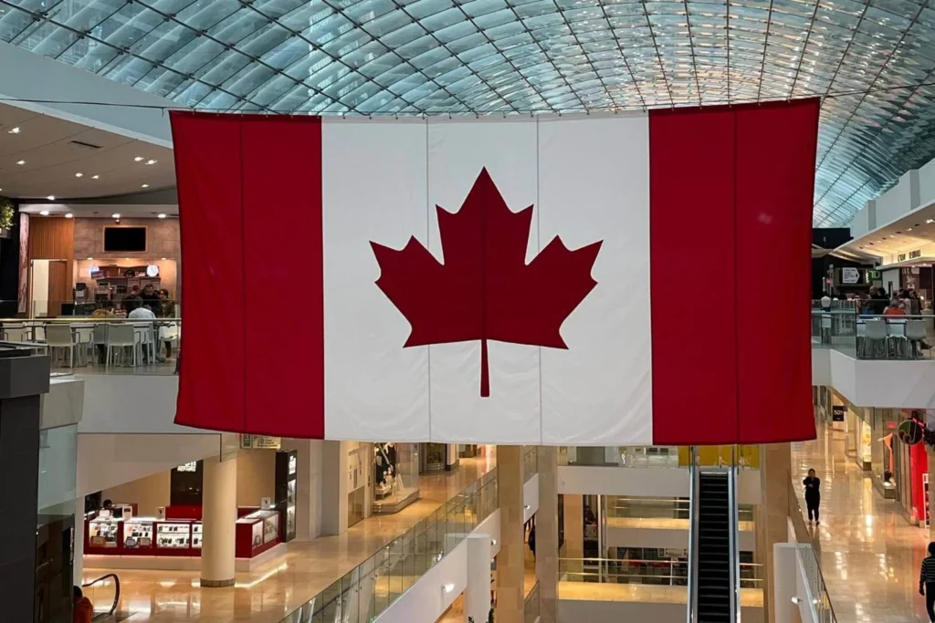 A massive Canadian flag hanging inside the Chinook Centre shopping mall in Calgary, with a curved glass ceiling above. Below, the mall's interior includes multiple levels of shops, a food court, and escalators. The red and white colors of the flag stand out prominently in the space