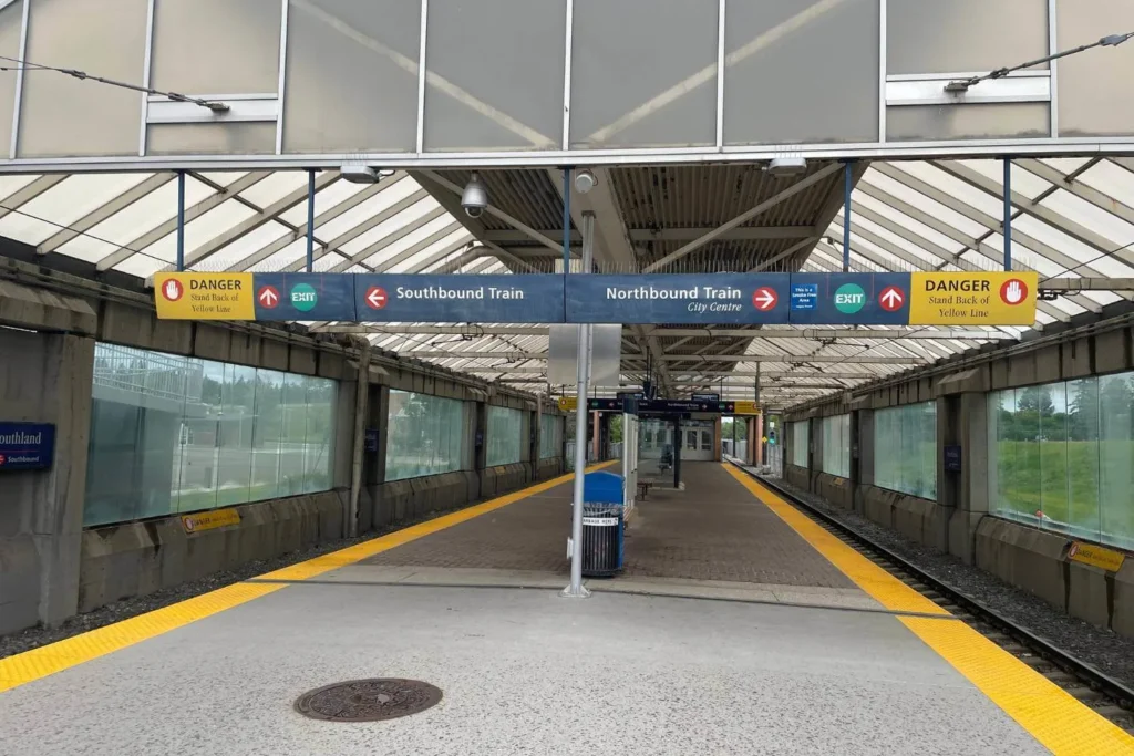 The Heritage CTrain Station in Calgary, an outdoor transit platform with a covered waiting area. A central sign directs passengers to the southbound and northbound trains, with additional safety warnings instructing people to stand behind the yellow line. The station has a modern, open design with glass-paneled walls, yellow tactile paving, and a trash bin near the entrance.