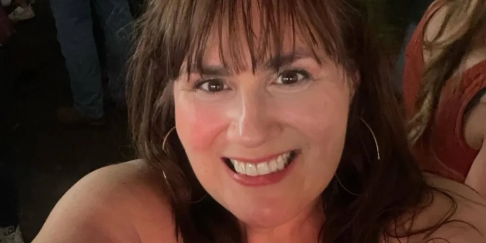 A woman with long brown hair and bangs takes a cheerful selfie in a crowded, dimly lit setting. She’s smiling brightly, wearing hoop earrings and sunglasses perched on her head, with people mingling in the blurry background.