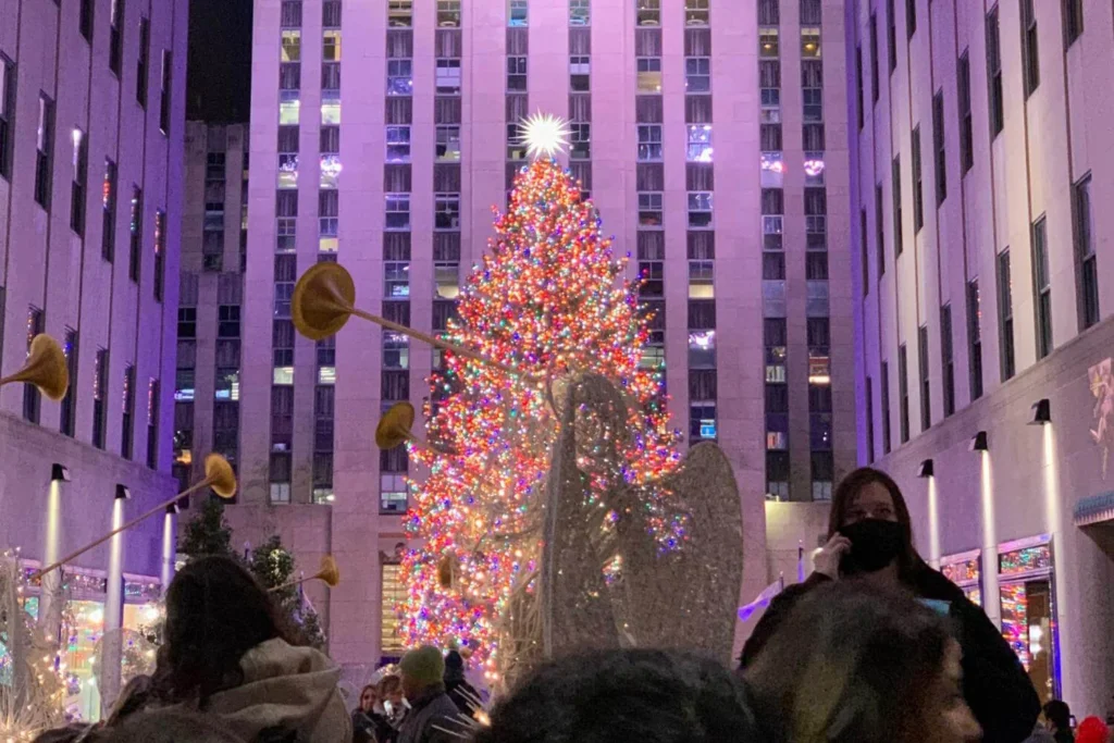 A tall Christmas tree glowing with colorful lights stands in the center of Rockefeller Plaza in New York City. The tree is surrounded by golden trumpeting angel decorations and crowds of bundled-up visitors taking in the festive nighttime scene.