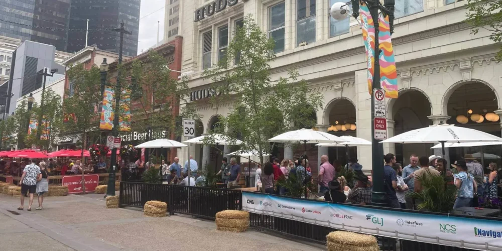 A lively street scene outside Hudson’s Bay in Calgary, with people gathered in a fenced-off outdoor patio under white umbrellas. Colorful banners hang from the lampposts, and red patio umbrellas add vibrancy to the scene. Skyscrapers in the background reflect the mix of historic and modern architecture.