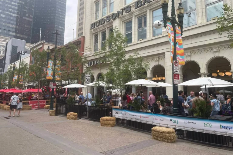 A lively street scene outside Hudson&rsquo;s Bay in Calgary, with people gathered in a fenced-off outdoor patio under white umbrellas. Colorful banners hang from the lampposts, and red patio umbrellas add vibrancy to the scene. Skyscrapers in the background reflect the mix of historic and modern architecture.