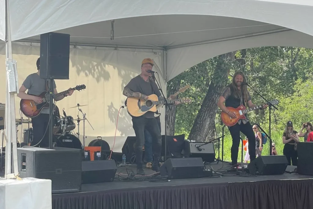 A live outdoor music performance under a white event tent. A three-member band is on stage, with a tattooed lead singer playing an acoustic guitar, a guitarist with long hair playing an electric guitar, and a partially visible drummer behind them. The audience watches from a grassy area, with some taking photos or enjoying the show in a relaxed, sunny setting in Calgary.