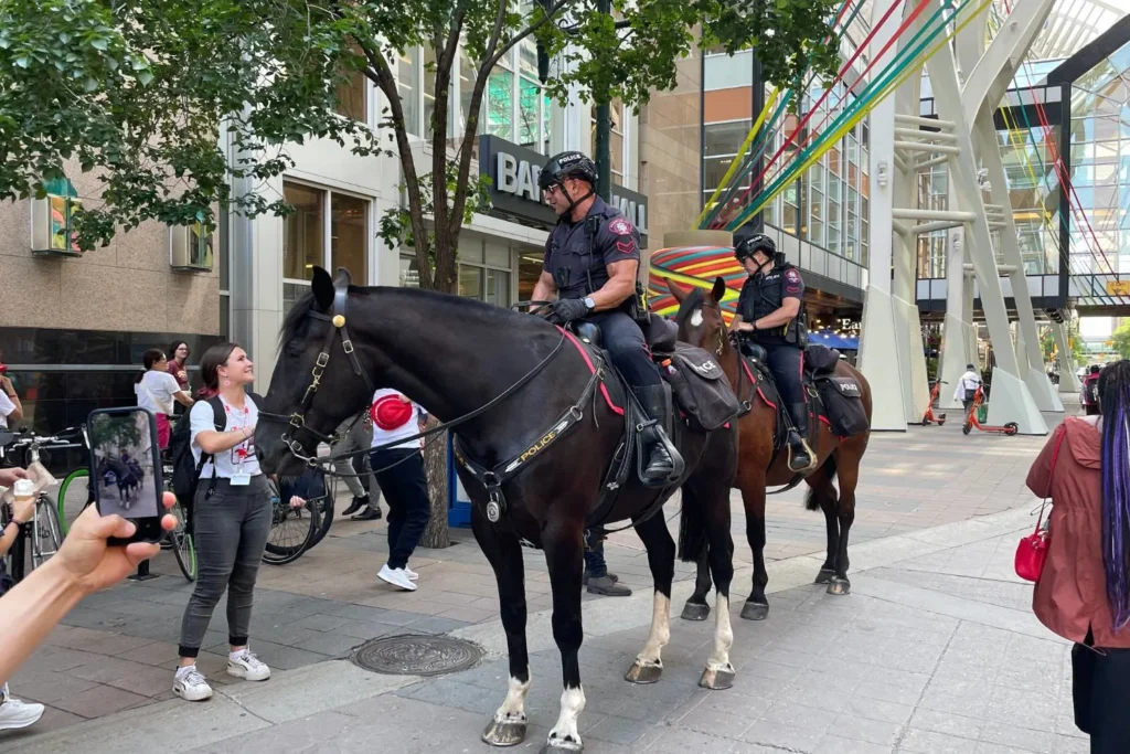 Two police officers in Calgary on horseback patrol a busy pedestrian street, engaging with the public. A woman in a white t-shirt smiles while petting one of the horses, as another person captures the moment on their phone. The background features modern buildings with colorful ribbon decorations, bicycles, and people walking or riding scooters.