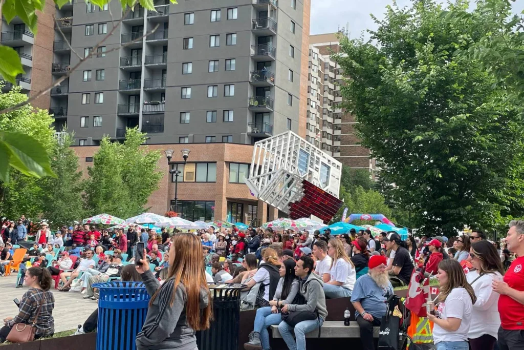 A large crowd gathers in an urban plaza in Calgary for an outdoor event, many wearing red and white, celebrating Canada Day. People sit on colorful chairs or stand under umbrellas, with some waving small Canadian flags. A striking, tilted cube-shaped public art installation stands in the background, along with tall residential buildings and lush green trees.