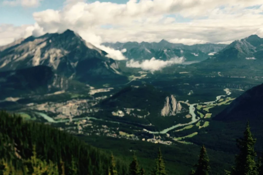 a breathtaking aerial view of a mountain valley in the Canadian Rockies. A towering, rugged mountain dominates the scene, with clouds partially wrapping around its peak. Below, a winding turquoise river snakes through the lush green valley, where a small town or village is nestled among the trees. The foreground features evergreen trees, adding depth to the expansive landscape.