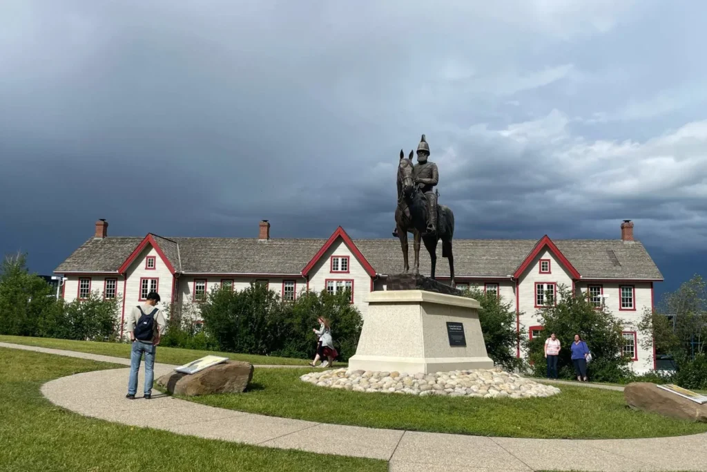 a bronze statue of a mounted police officer on a horse, standing on a stone pedestal in front of a historic white building with red trim. The Confluence historic site building has a classic design with multiple chimneys and a shingled roof, likely a heritage site or museum. A few visitors are walking along the curved pathway, reading plaques, and admiring the scene under a dramatic sky with dark storm clouds in the background.