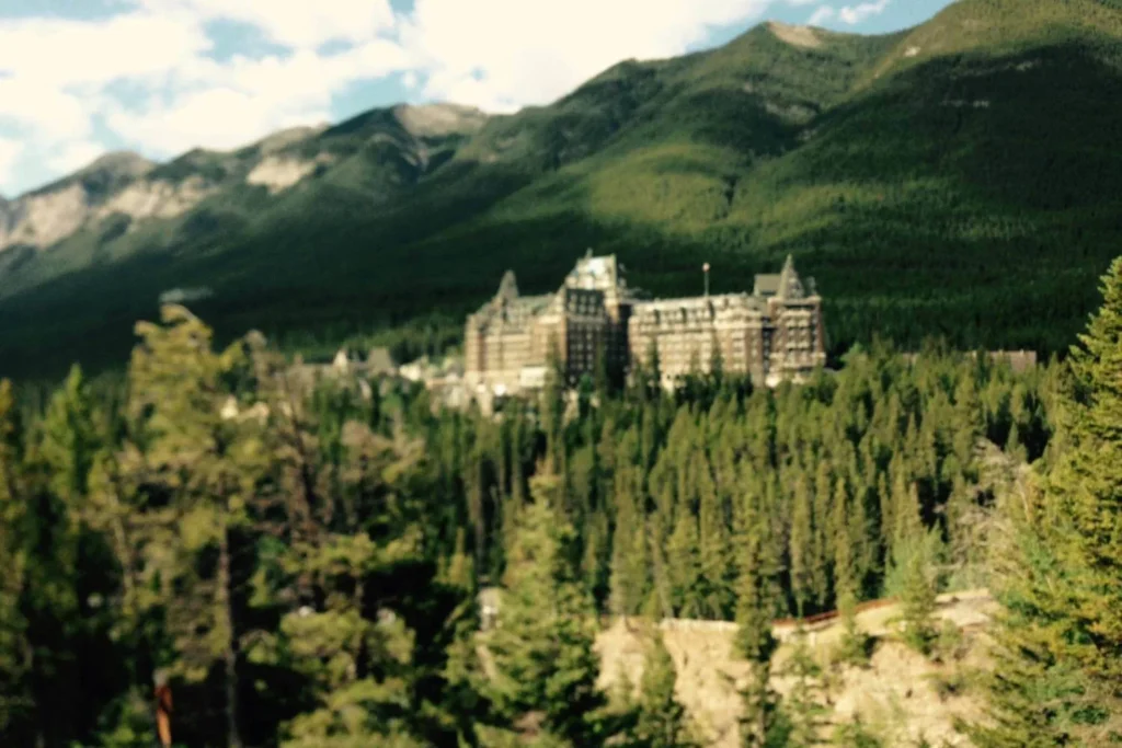 The Fairmont Banff Springs Hotel, a grand historic castle-like building nestled among dense evergreen forests in the Canadian Rockies. The hotel's iconic architecture stands out against the lush green mountain backdrop. Sunlight casts shadows on the slopes, adding depth to the breathtaking alpine scenery.