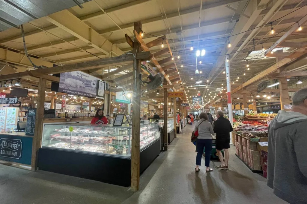 The vibrant indoor Calgary farmers' market with rustic wooden stalls selling fresh produce, meats, and specialty goods. A butcher counter labeled "Spragg's Meat Shop" displays various cuts of pork, with a vendor in a red hoodie behind the glass case. Warm string lights hanging from the ceiling create a cozy atmosphere as shoppers browse the aisles filled with fruits, vegetables, and local delicacies.