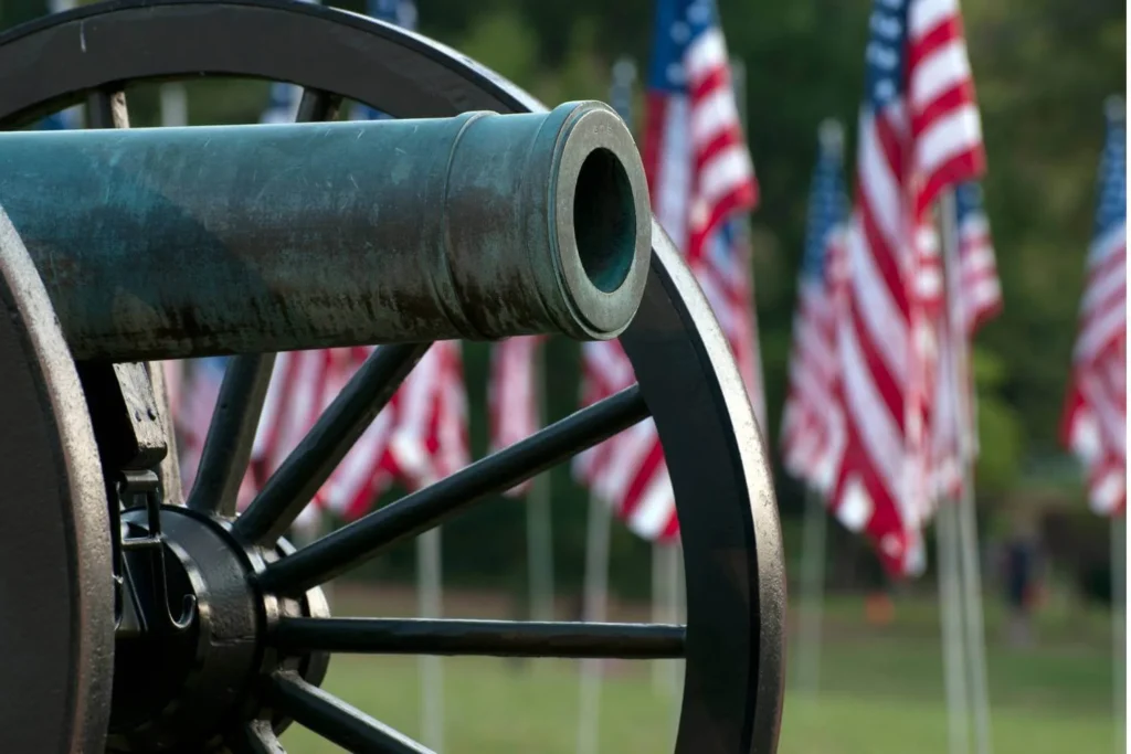 Close-up of an old bronze cannon wheel and barrel pointing slightly upward, with a field of blurred American flags waving in the background. The image captures a patriotic and historic tone with natural greenery beyond the flags.