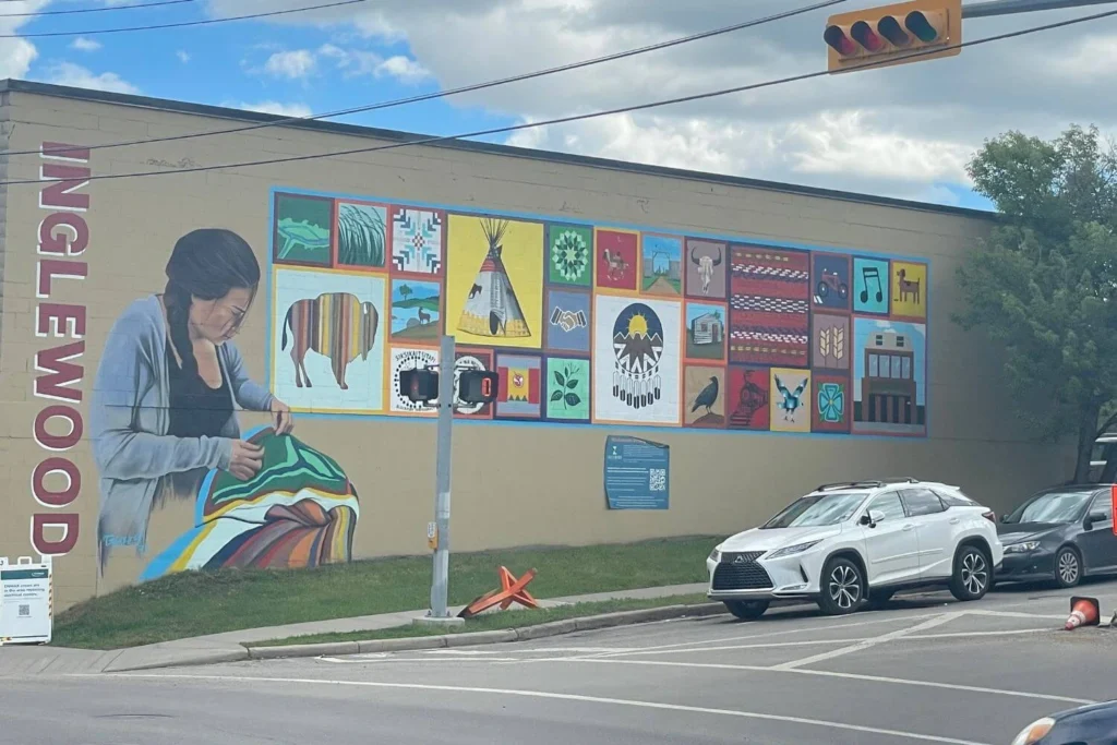 Large mural on the side of a building in Inglewood showing a woman with braided hair sewing a colorful blanket, surrounded by a vibrant patchwork of Indigenous-inspired artwork including a bison, tipi, handshake, dreamcatcher, and nature motifs.