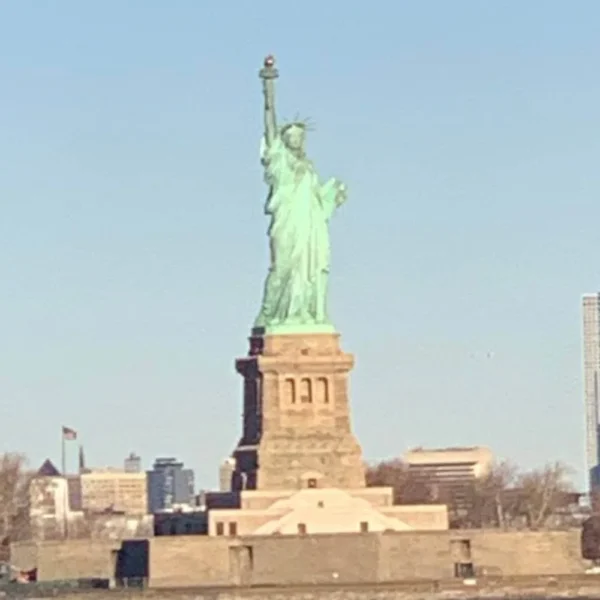 Distant view of the Statue of Liberty standing tall on Liberty Island with a clear blue sky behind it and modern skyscrapers visible in the background. The statue's green patina contrasts with the stone pedestal and surrounding bare trees.